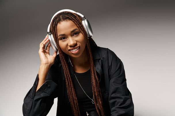 pleased african american woman in 20s with dreadlocks listening music in headphones in grey studio - Photo, Image