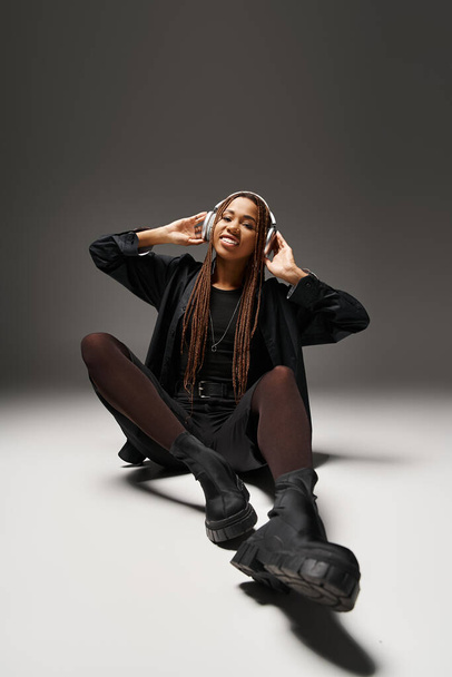 sonriente mujer afroamericana de 20 años con rastas escuchando música en auriculares en estudio gris - Foto, imagen