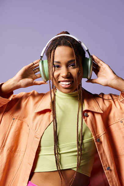 chica afroamericana feliz en los años 20 con auriculares y atuendo casual vibrante sobre fondo púrpura - Foto, Imagen
