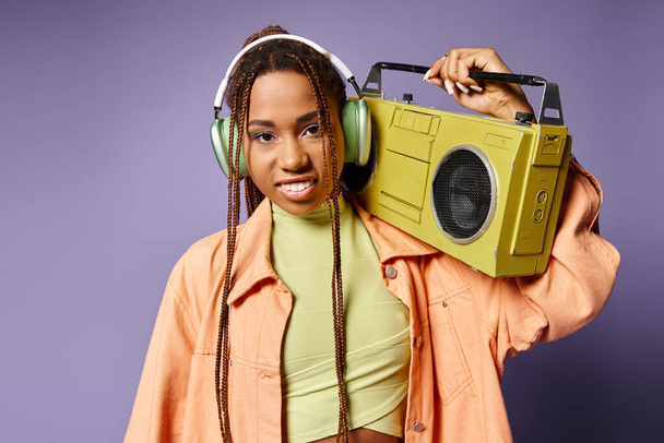 紫色の背景にレトロブームボックスで立っているワイヤレスヘッドフォンの幸せなアフリカ系アメリカ人女性 - 写真・画像