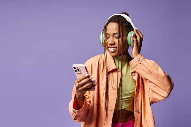 glückliche afrikanisch-amerikanische Frau, die Musik in ihren Kopfhörern hört und das Telefon auf fliederfarbenem Hintergrund benutzt - Foto, Bild