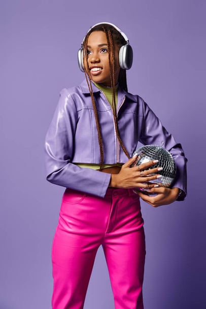 улыбающаяся молодая африканская американка в стильной одежде и наушниках, держащая диско-мяч на фиолетовом - Фото, изображение