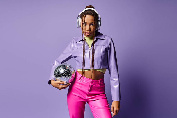 joven afroamericana mujer en traje de moda y auriculares sosteniendo bola disco sobre fondo púrpura - Foto, imagen