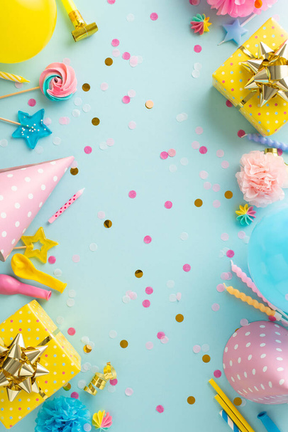 Joyful festivity creativity. Vertical top view of table set with sweets, wrapped gifts, party hats, noise-maker, candles, straws, balloons, confetti on soft blue backdrop with available space for text - Photo, Image