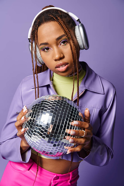 κομψό αφρικανικό αμερικανικό κορίτσι σε μοντέρνα ασύρματα ακουστικά κρατώντας ντίσκο μπάλα σε μωβ φόντο - Φωτογραφία, εικόνα