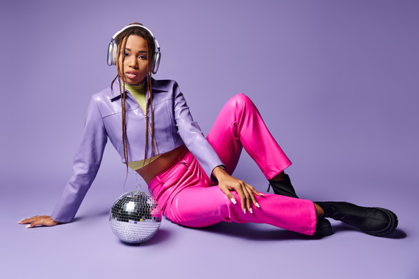 chica afroamericana con estilo en auriculares inalámbricos sentados cerca de la bola disco en el telón de fondo púrpura - Foto, Imagen