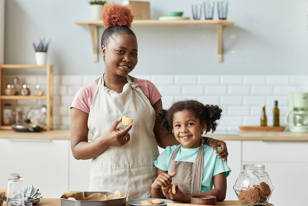 Portrait à la taille d'une mère et d'une fille heureuses dégustant des biscuits frais faits maison et regardant la caméra tout en appréciant de cuisiner ensemble dans la cuisine - Photo, image