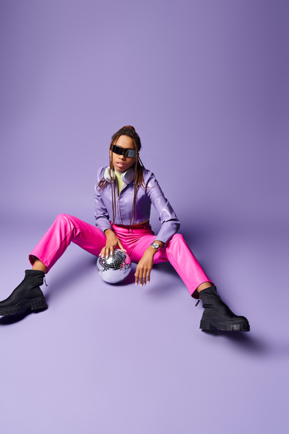 stylish african american girl in trendy sunglasses and headphones sitting near disco ball on purple - Zdjęcie, obraz