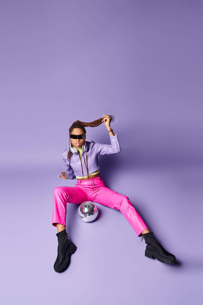 mujer afroamericana en gafas de sol futuristas y auriculares sentados cerca de la bola disco en púrpura - Foto, imagen