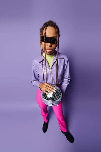 young african american woman in trendy sunglasses holding disco ball on chain and standing on purple - Photo, Image