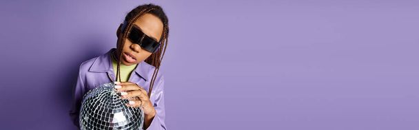 pancarta de mujer afroamericana en gafas de sol de moda sosteniendo bola disco sobre fondo púrpura - Foto, Imagen