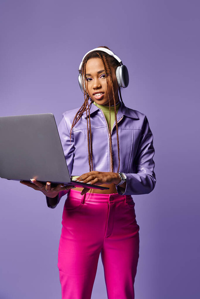 紫色の背景でリモートでラップトップに取り組んでいるヘッドフォンを持つ若いアフリカ系アメリカ人女性 - 写真・画像