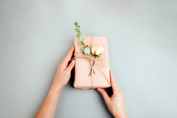 Background for greetings. Gift wrapping in soft pink paper with dry eucalyptus branch on a light concrete background in womens hands. Top view - Photo, Image