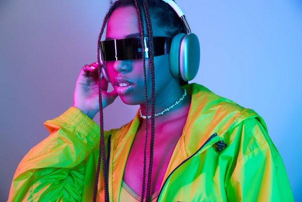young african american girl in wireless headphones and jacket standing in studio with neon lights - Photo, Image