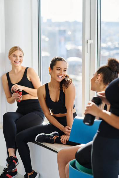 alegre interracial jóvenes mujeres en 20s charlando mientras sentado al lado de ventana en yoga estudio - Foto, Imagen