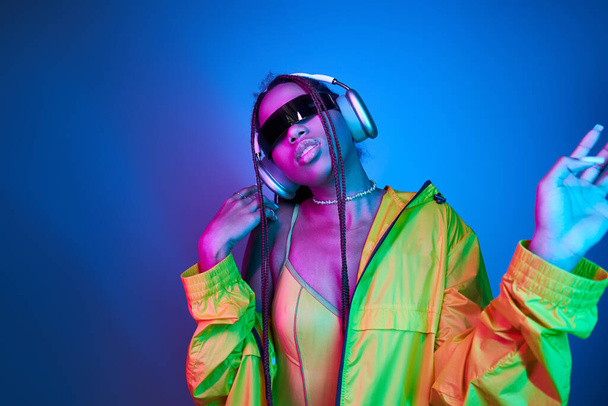 afro-américaine fille dans des écouteurs et des lunettes de soleil élégantes écouter de la musique en studio avec des lumières - Photo, image