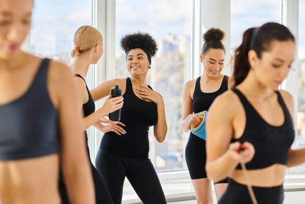 cinq jeunes femmes multiculturelles en tenue active bavardant et souriant après l'entraînement en studio pilates - Photo, image
