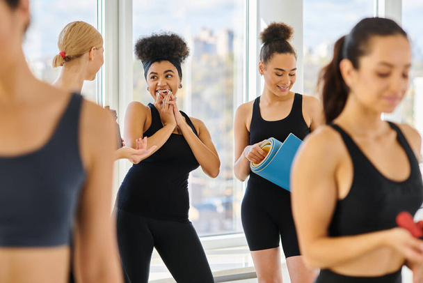 cinco mujeres felices y jóvenes multiculturales en activo desgaste charlando después del entrenamiento en el estudio de pilates - Foto, imagen