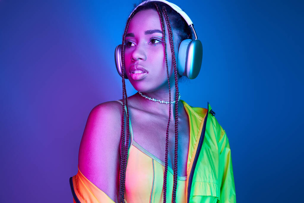 Nachdenkliche dunkelhäutige Frau mit drahtlosen Kopfhörern posiert in Jacke im Studio mit Neonlicht - Foto, Bild