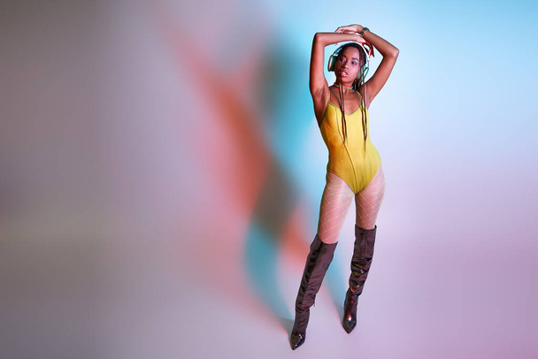 young dark-skinned woman in headphones posing in bodysuit and over knee boots in studio with lights - Photo, Image