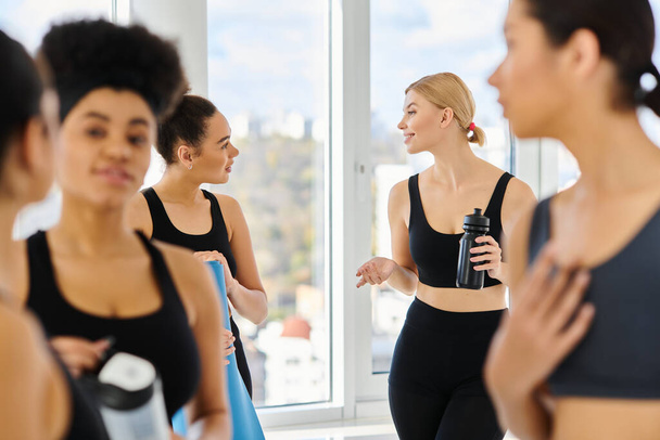 happy blonde woman holding sports bottle and talking with her african american friend in yoga studio - Photo, Image