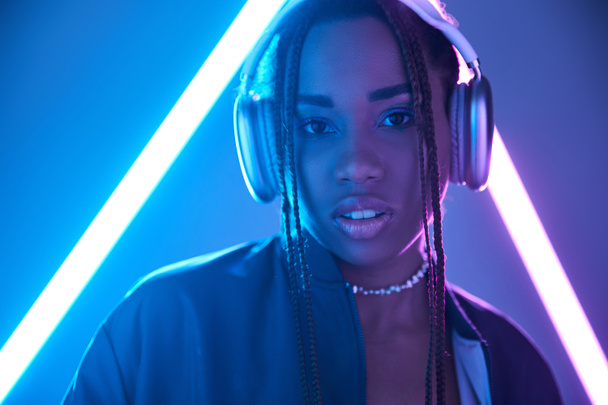 portrait of african american girl in dreadlocks and headphones in studio with blue fluorescent light - Photo, Image