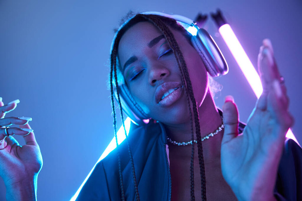 ブルー蛍光灯でスタジオで音楽を楽しんでいるヘッドフォンの若いアフリカ系アメリカ人女性 - 写真・画像