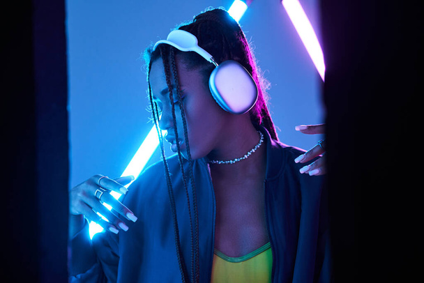довольно африканская американка в наушниках наслаждается музыкой в студии с синим флуоресцентным светом - Фото, изображение