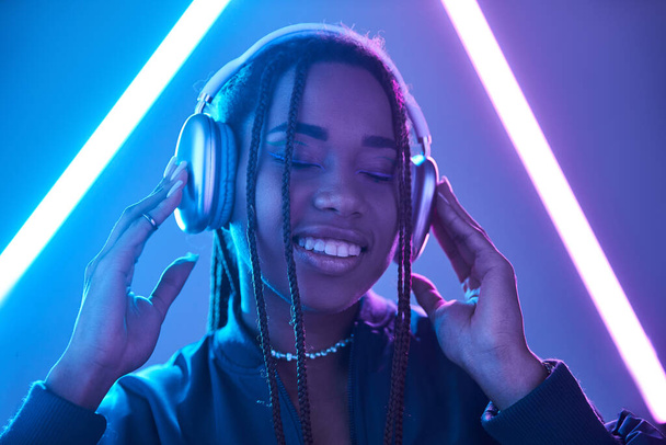 χαρούμενη Αφροαμερικανή γυναίκα με ακουστικά που απολαμβάνει μουσική στο στούντιο με φθορίζον φως - Φωτογραφία, εικόνα