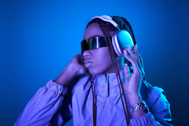 bonita mujer afroamericana posando en gafas de sol de moda con auriculares en azul con luz de neón - Foto, Imagen