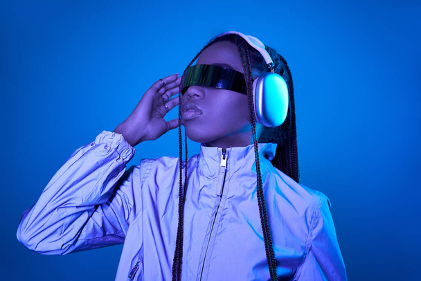 красива темношкіра жінка позує в модних сонцезахисних окулярах з навушниками на синьому з неоновим світлом - Фото, зображення