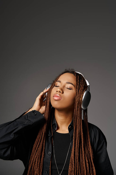 junge dunkelhäutige Frau mit Dreadlocks genießt Musik in drahtlosen Kopfhörern vor grauem Hintergrund - Foto, Bild