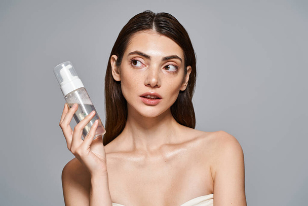 Μια νεαρή λευκή γυναίκα με καστανά μαλλιά κρατά ένα μπουκάλι με αφρό καθαρισμού προσώπου μπροστά από το πρόσωπό της, προβάλλοντας καθαρό δέρμα. - Φωτογραφία, εικόνα