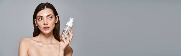 Młoda kaukaska kobieta z brunetką z włosami wygląda na zaskoczoną trzymając butelkę środka do czyszczenia twarzy w studio, sztandar - Zdjęcie, obraz