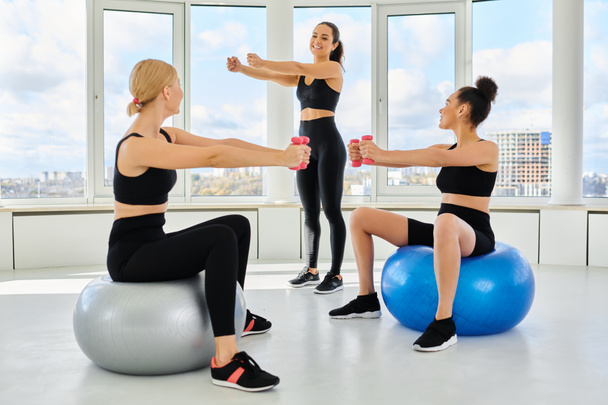 alegres amigas multiculturales haciendo ejercicio con pesas en las pelotas de fitness cerca del entrenador - Foto, imagen