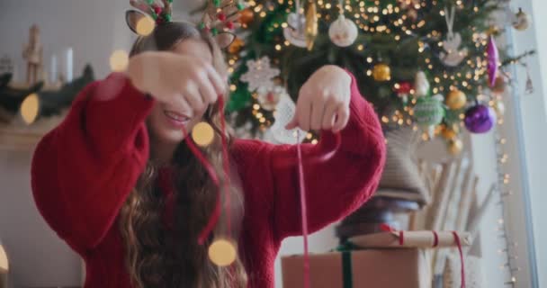 Hermosa mujer joven usando cinta roja para envolver regalos con árbol de Navidad decorado en el fondo en casa - Metraje, vídeo