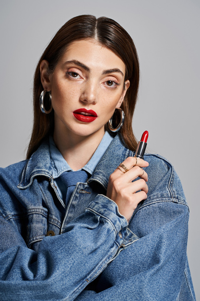 A young caucasian woman in a jean jacket happily holds a lipstick, embodying style and elegance. - Photo, Image