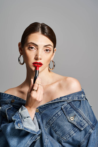 A young Caucasian woman with brunette hair wearing a jean jacket, holding a vibrant red lipstick. - Photo, Image
