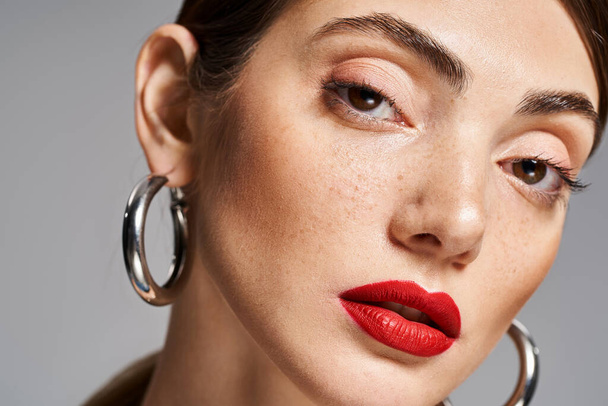 A young Caucasian woman with brunette hair sporting red lipstick and hoop earrings exudes confidence and style. - Photo, Image