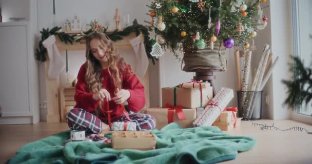 Щаслива молода жінка зав'язує стрічку на загорнутий різдвяний подарунок, сидячи на ковдрі вдома - Кадри, відео