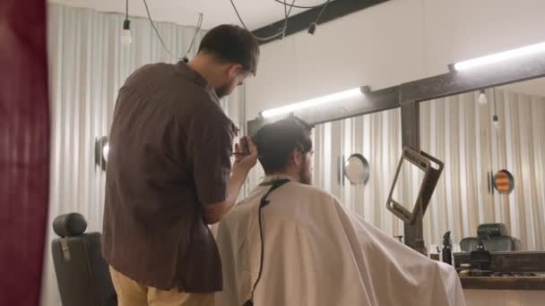 Hombre cliente sentado frente al espejo en la barbería mientras que el peluquero profesional le da un nuevo corte de pelo - Imágenes, Vídeo