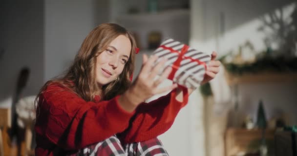 Szczęśliwa piękna młoda kobieta wiruje opakowane pudełko prezentów siedząc w jasnym domu podczas Bożego Narodzenia - Materiał filmowy, wideo