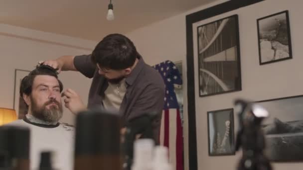 Szakállas férfi fodrász köpenyben ül a fodrászatban, miközben fiatal borbély vágja a haját, amerikai zászló és fekete-fehér fotók a falon a háttérben - Felvétel, videó
