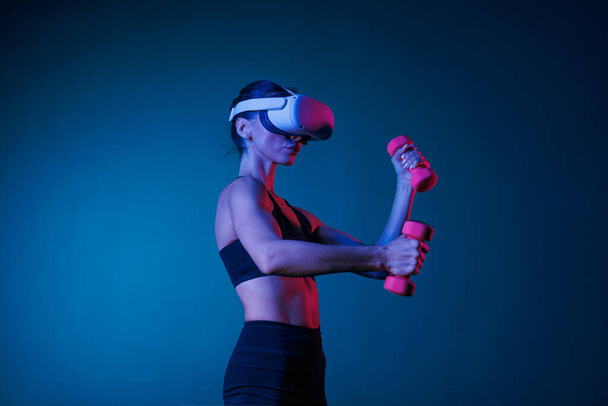 Sportliches Mädchen beim Online-Training in VR-Brille in schwarzer Kleidung mit Hanteln auf dunklem Hintergrund, Online-Sporttraining - Foto, Bild