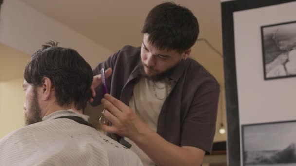 Молодий професійний перукар для стрижки волосся людини з ножицями і гребінцем під час робочого дня в перукарні - Кадри, відео