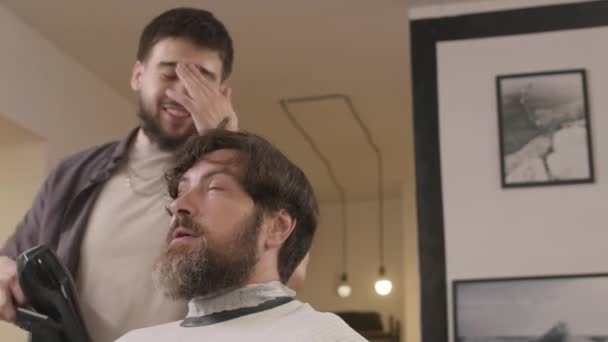 Vidám borbély beszélgetés férfi ügyféllel, miközben szárítja a haját hajvágás után a fodrászatban - Felvétel, videó