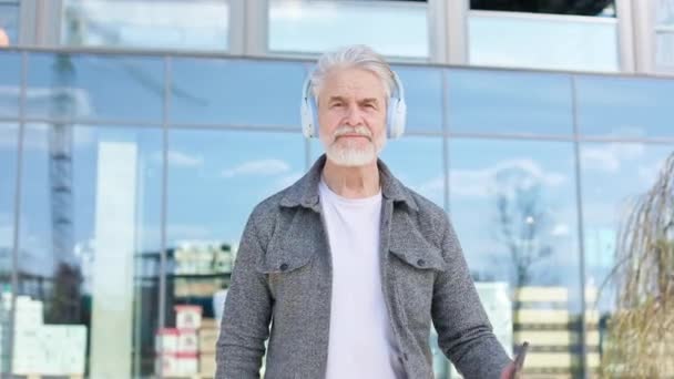 Portret pewnego siebie przystojnego emeryta stojącego w pobliżu odwzorowanego domu w centrum miasta. Kaukaski siwowłosy mężczyzna w słuchawkach spędza czas w nowoczesnej miejskiej atmosferze w słoneczny dzień. - Materiał filmowy, wideo