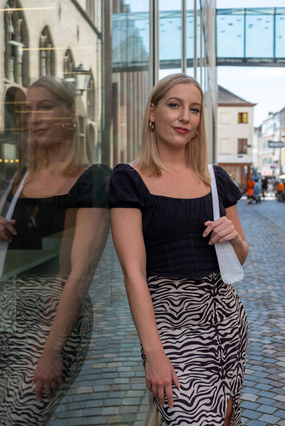 Dieses Bild zeigt eine junge kaukasische Frau mit blonden Haaren, die eine Mischung aus Ehrgeiz und Stil widerspiegelt, während sie vor einem gläsernen Gebäude in einer städtischen Umgebung steht. Ihre Spiegelung auf der Glasoberfläche - Foto, Bild