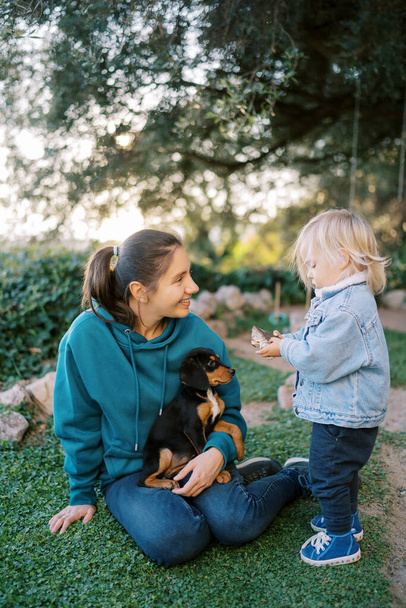 Klein meisje staat naast een lachende moeder zittend met een puppy op haar knieën op het gazon. Hoge kwaliteit foto - Foto, afbeelding