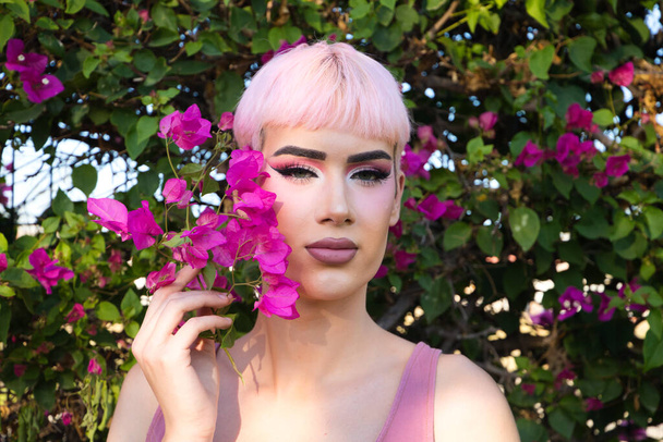 Retrato de un joven gay con el pelo y el maquillaje de color rosa tiene al lado de su cara una flor de buganvilla púrpura. El chico hace diferentes expresiones. Concepto de igualdad y derechos LGBTQ - Foto, imagen